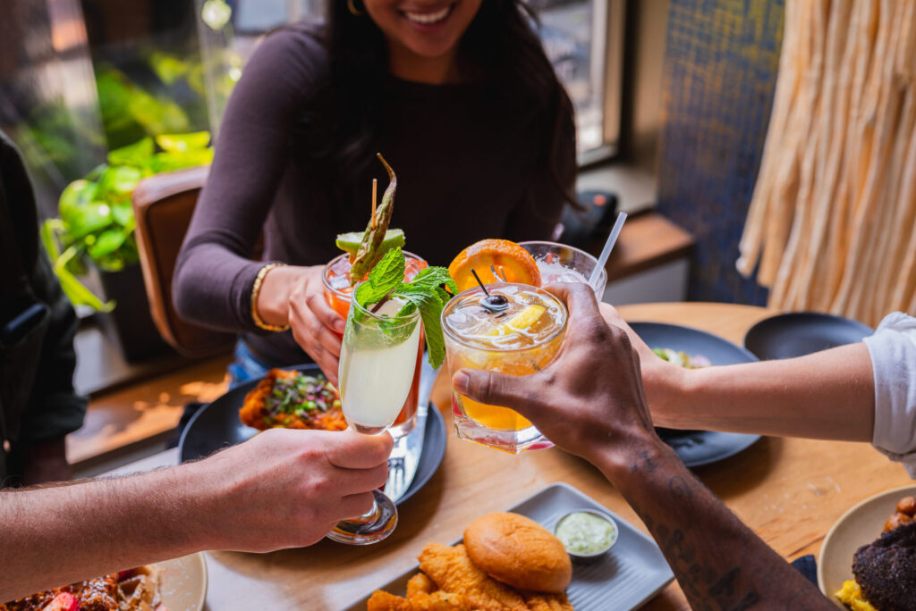 Four people toast with different cocktails over a table filled with food, including fried chicken, a sandwich, dipping sauce, and pasta. Sunlight streams in, creating a cheerful, lively fall atmosphere.
