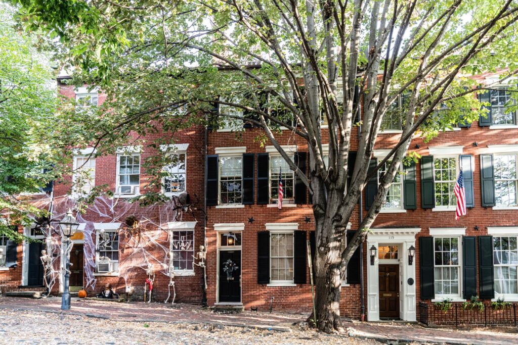 A street with brick row houses decorated for Halloween in the fall, featuring white fake spider webs, skeletons, pumpkins, and American flags, with a large tree shading the sidewalk and fallen leaves scattered on the ground.