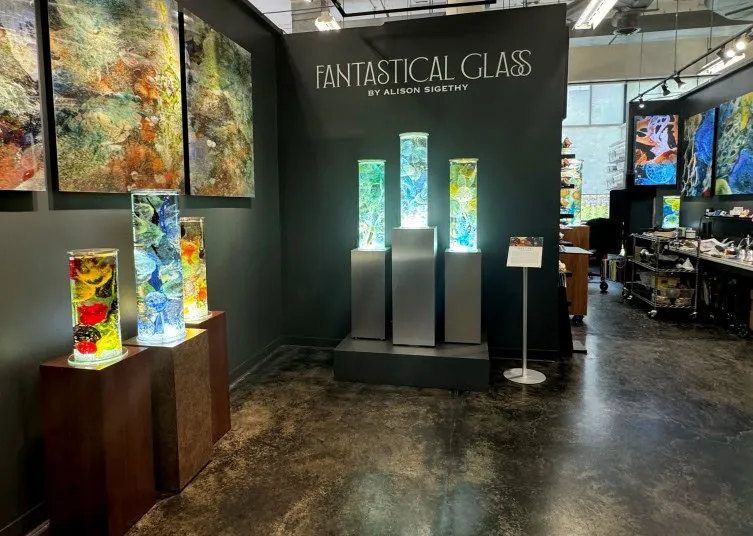 An art gallery display titled FANTASTICAL GLASS BY ALISON SIGETHY features colorful glass sculptures in tall cylindrical cases, surrounded by vibrant abstract wall art. The gallery has dark walls and polished floors.