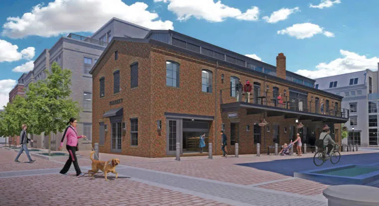 A brick building with large windows and a second-floor terrace. People walk on the sidewalk, including a woman with a dog, and others cycle nearby. Trees and modern buildings are in the background under a blue sky with clouds.