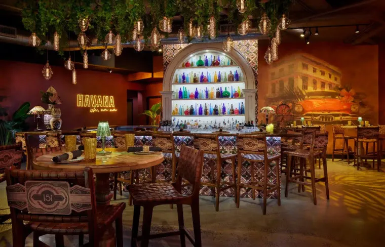 A vibrant Cuban-themed bar with colorful bottles on a backlit arched shelf, ornate chairs, hanging lights, greenery, and warm decor. HAVANA glows on the wall, with a mural of a vintage car and building in the background.