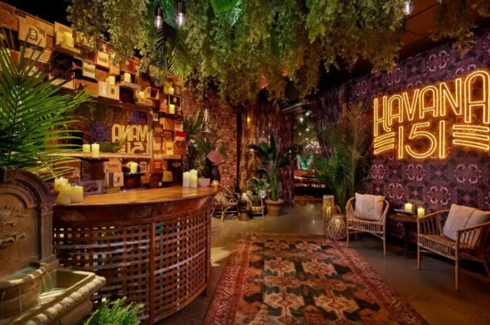 A cozy, tropical-themed lounge with lush hanging greenery, vintage decor, wicker chairs, glowing candles, a wooden bar, and a neon Havana 151 sign on a patterned wall. Rug and palm plants complete the warm atmosphere.