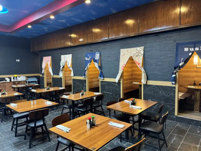 A modern Japanese restaurant with wooden tables and chairs. Cozy booth seating areas with fabric curtains line one wall. Soy sauce bottles and chopsticks are set on the tables, and the overall ambiance is warm and inviting.