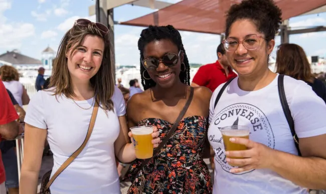 Three women smile at the camera while holding drinks at an outdoor event. One wears a white t-shirt, another a floral strapless dress, and the third a white Converse shirt. Other people and sunny skies are visible in the background.