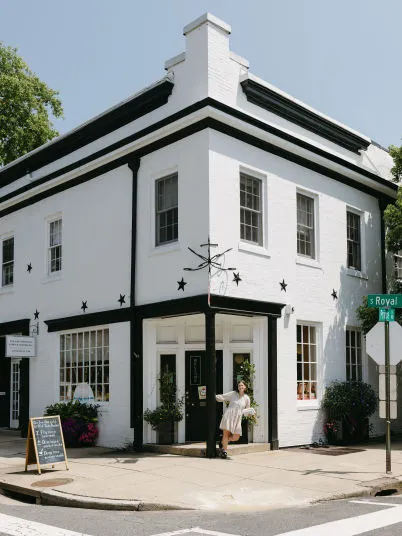 A woman stands in front of a white, two-story corner building decorated with black trim and star accents on a sunny day. There are potted plants, sidewalk signs, and street signs for Royal St. and Wilkes St. visible.