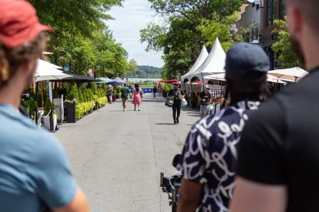Three people in the foreground watch as others walk down a tree-lined street with outdoor seating, market umbrellas, and tents on a sunny day. The scene is lively with greenery and people enjoying the area.