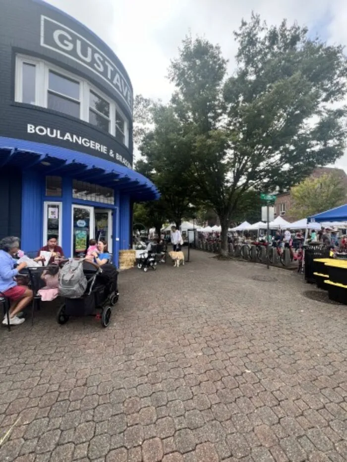 People sit at outdoor tables near a blue bakery frontage on a tree-lined brick plaza, while others walk or stand in line for vendor tents at a street market in the background.