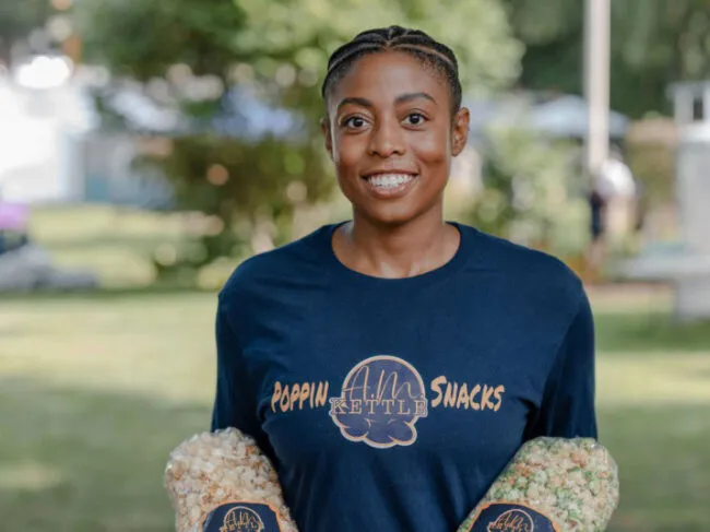 A smiling person wearing a navy “A.M. Kettle Poppin Snacks” shirt holds two bags of colorful popcorn outdoors with trees and blurred greenery in the background.