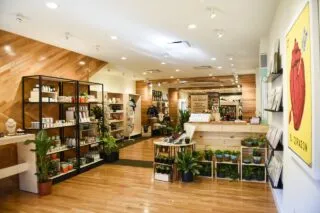 A modern boutique shop with wooden floors and walls, shelves displaying plants, pots, and various goods, and a large yellow artwork with a red anatomical heart on the right wall. The space is brightly lit and inviting.