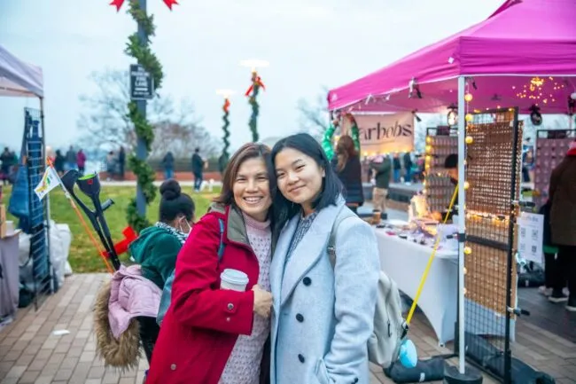 Two women smiling and standing close together at an outdoor market, with festive decorations, vendor tents, and people in the background. One wears a red coat and holds a coffee cup; the other wears a light gray coat.
