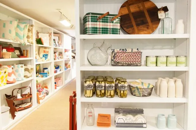 A white shelving unit displays jars of pickles, milk bottles, canned goods, glass pitchers, and kitchen items, with more colorful gifts, bags, and home décor on shelves in the background.