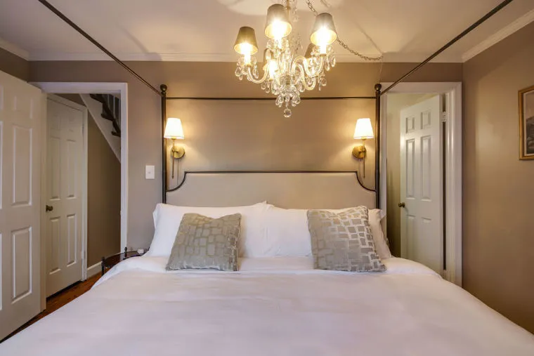 A cozy bedroom with a large bed, white bedding, two decorative pillows, wall-mounted lamps, and a chandelier. The walls are beige, and there are open doors on either side of the room.