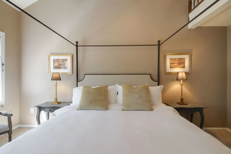 A neatly made bed with white bedding and beige pillows is centered between two bedside tables, each with a lamp and a framed picture above, in a softly lit, neutral-toned bedroom.