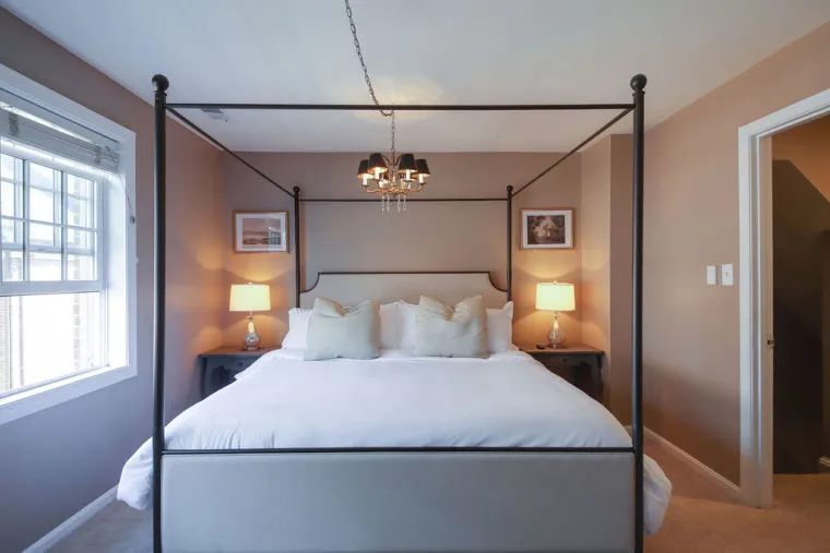 A bedroom with a four-poster bed, white bedding, two bedside tables with lamps, framed pictures on the taupe walls, a window on the left, and a small chandelier hanging from the ceiling.