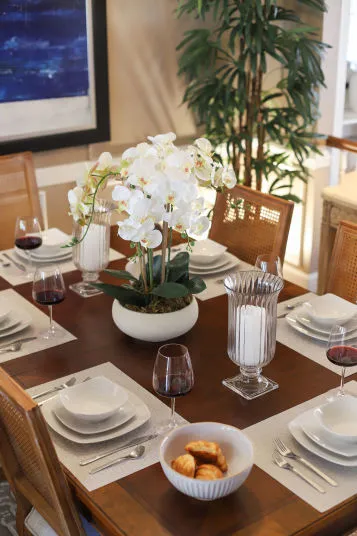 A dining table set for four with white dishes, wine glasses with red wine, and a centerpiece of white orchids. A bowl of pastries is in the middle. A tall green plant and framed artwork are in the background.