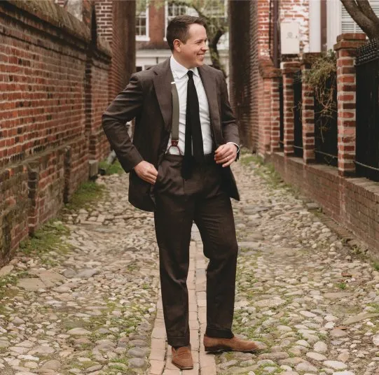 A man in a brown suit with suspenders and a tie stands on a cobblestone alley, smiling and looking to the side. Brick walls and small trees line the narrow walkway.