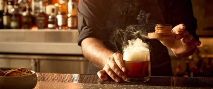 A bartender uses a smoking device to infuse a cocktail with smoke at a bar, creating a dramatic smoky effect above the glass. Bottles of liquor are visible blurred in the background.