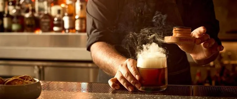 A bartender uses a smoking device to infuse a cocktail with smoke at a bar, creating a dramatic smoky effect above the glass. Bottles of liquor are visible blurred in the background.