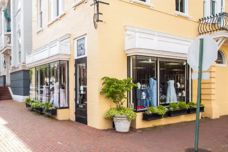 A clothing boutique with large display windows on a street corner, featuring dresses and outfits inside. The exterior is light yellow brick with white trim, and planters with green plants line the windows.