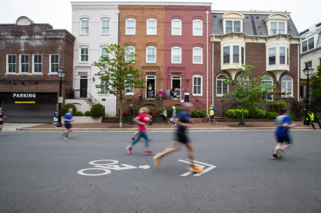 People jogging and walking on a street in front of colorful row houses. The street has a bike lane symbol, and the image is slightly blurred, indicating motion. Some onlookers stand on the steps of the houses.