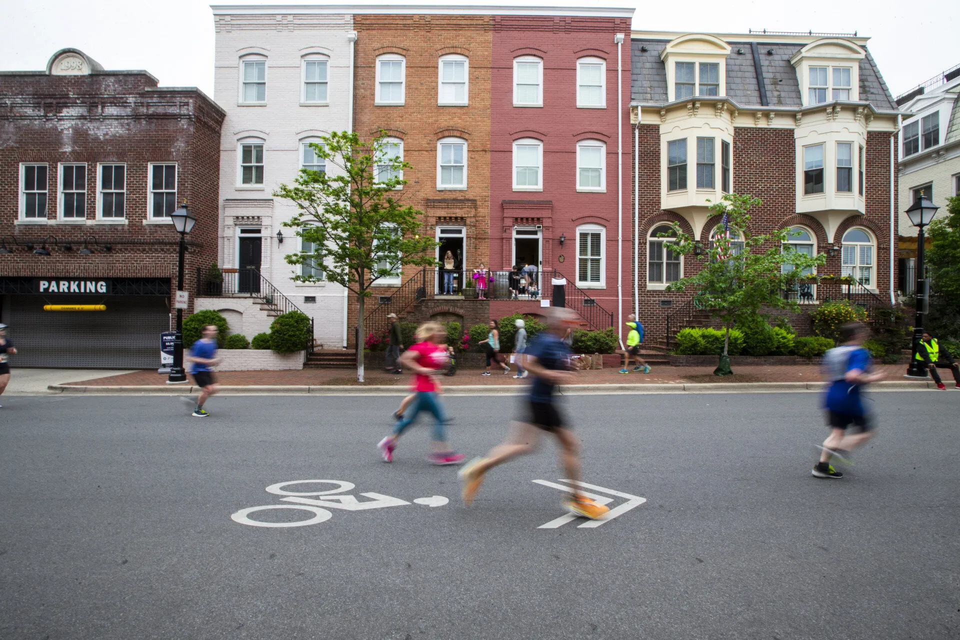 People jogging and walking on a street in front of colorful row houses. The street has a bike lane symbol, and the image is slightly blurred, indicating motion. Some onlookers stand on the steps of the houses.