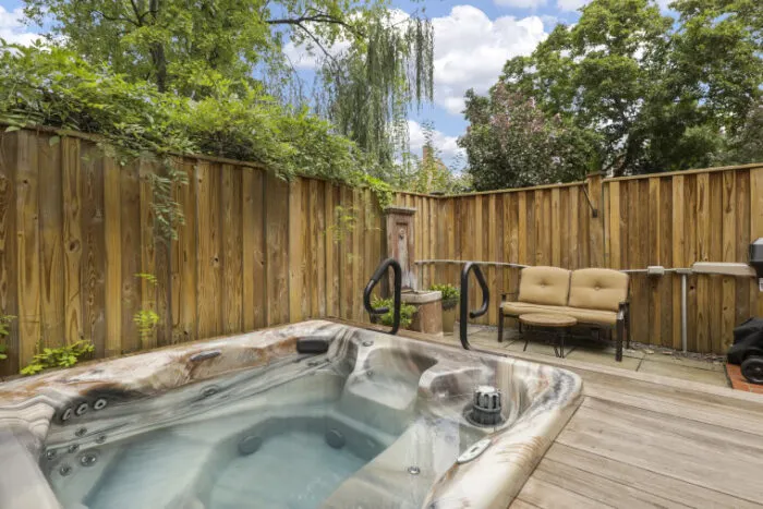 A backyard deck with a hot tub, small fountain, tan cushioned bench, wooden privacy fence, surrounded by lush greenery and trees under a partly cloudy sky.