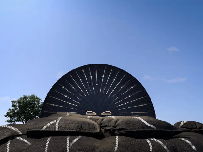 A large decorative black fan with white patterns stands behind a pile of black sandbags with white stripes, under a clear blue sky with a small tree visible to the left.
