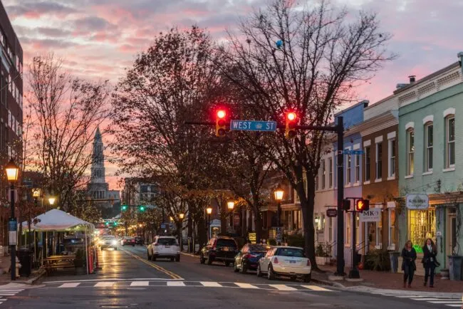 A charming city street at sunset with red traffic lights, parked cars, shops, leafless trees, and a distant clock tower under a colorful sky. Pedestrians walk on the sidewalk and street lamps light the scene.