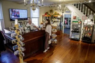 A woman stands at a wooden counter speaking with a man behind it in a quaint store with wooden floors, shelves of goods, a postcard rack, and a TV screen displaying a welcome message. Stairs and another room are visible in the background.