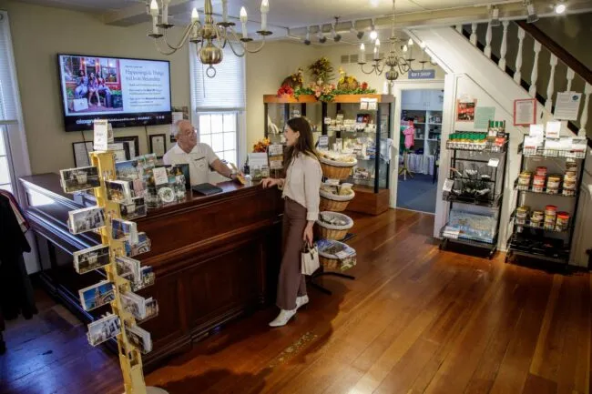 A woman stands at a wooden counter speaking with a man behind it in a quaint store with wooden floors, shelves of goods, a postcard rack, and a TV screen displaying a welcome message. Stairs and another room are visible in the background.