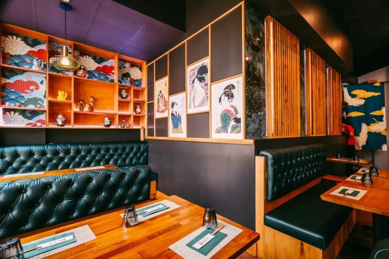 A cozy restaurant interior with teal tufted booths, wooden tables, Japanese art on the walls, decorative shelves, and colorful patterned wallpaper with a modern and inviting atmosphere.
