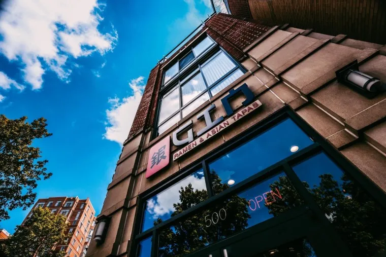 Low-angle view of a modern building with a restaurant sign reading Ramen & Asian Tapas above the entrance, a glass door showing 500 OPEN, and trees and blue sky with clouds in the background.