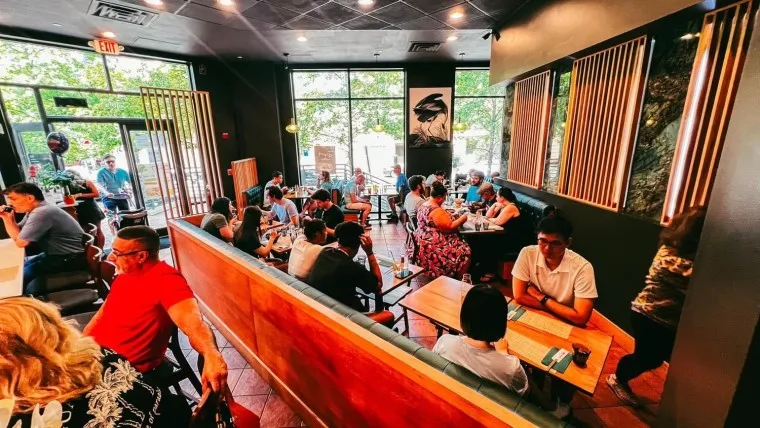 A bustling café filled with people sitting at tables and booths, chatting and dining. Large windows reveal trees and more patrons outside. Warm lighting and modern decor create a lively atmosphere.