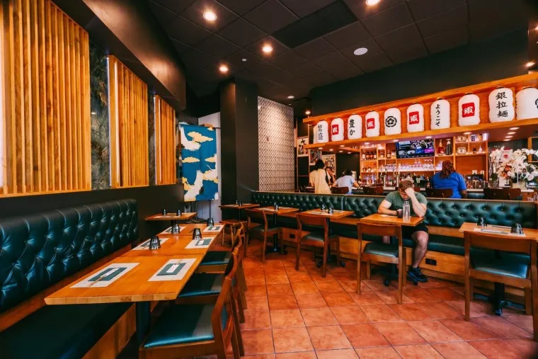 A modern Japanese restaurant interior with wooden tables, green cushioned benches, Japanese lanterns above the bar, and a few people seated. Warm lighting and decorative wall art create a cozy atmosphere.