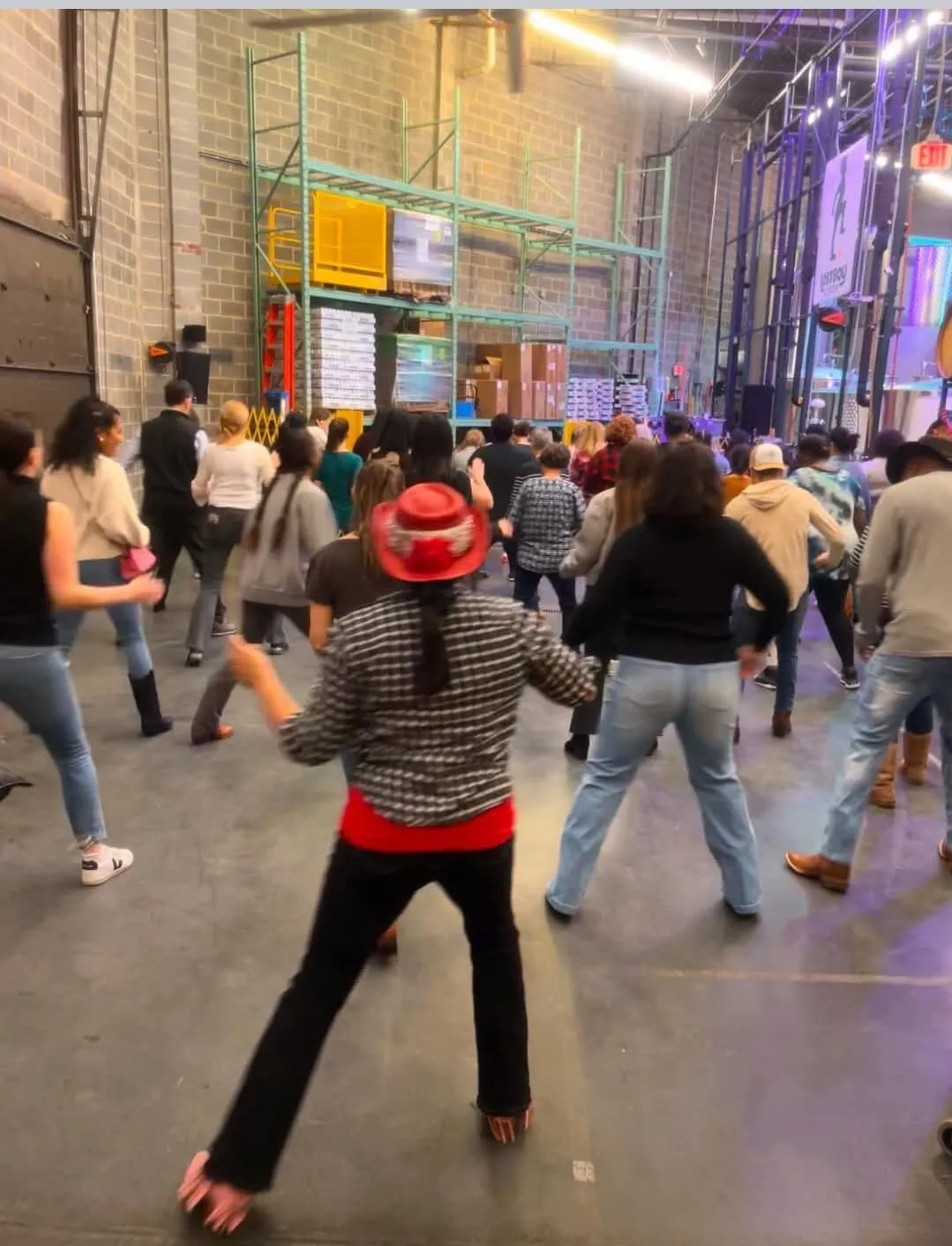 A group of people in casual clothes practice dance moves in a large industrial warehouse; a person in front wears a bright red hat and leads the group. Shelving and equipment are visible in the background.