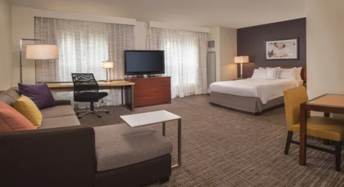 A spacious hotel room with a bed, sofa, desk with office chair, TV on a dresser, small dining table with two chairs, lamps, and large windows with sheer curtains.