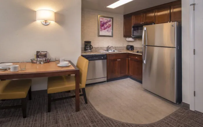 A modern hotel kitchenette with stainless steel appliances, wooden cabinets, a sink, and a countertop. A small dining table with two yellow chairs and place settings is beside the kitchen area.