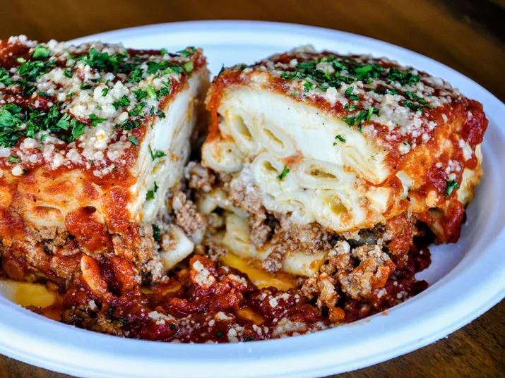 A close-up of a baked ziti pasta dish topped with tomato sauce, ground meat, melted cheese, and chopped parsley, served on a white plate with a portion cut to show the pasta and filling inside.