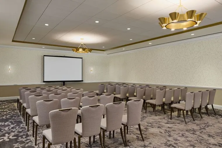 A conference room with rows of beige chairs facing a projection screen, patterned carpet, cream-colored walls, and two modern chandeliers hanging from the ceiling.