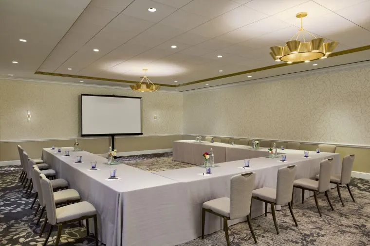 A conference room set up in a U-shape with beige chairs and tables draped in white cloths, each place set with a notepad, pen, bottled water, and a small flower vase. Two gold chandeliers and a projector screen are visible.