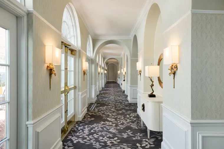A bright, elegant hallway with arched doorways, large windows, decorative wall sconces, patterned carpet, and white paneled walls. A console table with a lamp and mirror is positioned along the right wall.