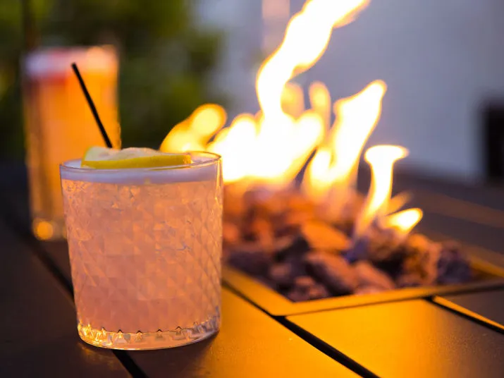 A close-up of a textured glass with a light cocktail, garnished with a lemon slice and a straw, on a dark table near a rectangular fire pit with visible flames. Another drink is blurred in the background.