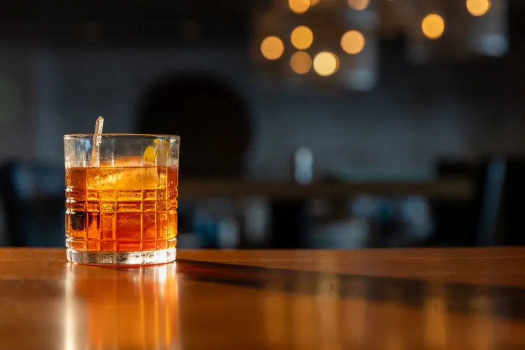 A glass of amber-colored cocktail with ice and an orange peel garnish sits on a wooden bar, with warm, blurry lights and a dimly lit background.