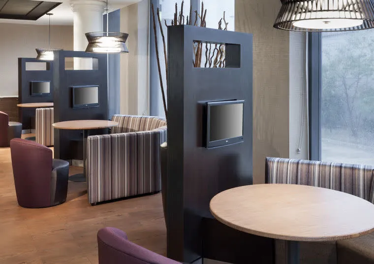 Modern lounge area with round tables, striped upholstered booths, purple chairs, and privacy dividers with mounted flat-screen TVs, next to large windows allowing natural light.