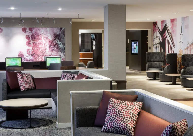Modern hotel lounge with gray and white seating, colorful patterned pillows, round tables, computer workstations, and abstract wall art in shades of pink and purple. The space is brightly lit and open.