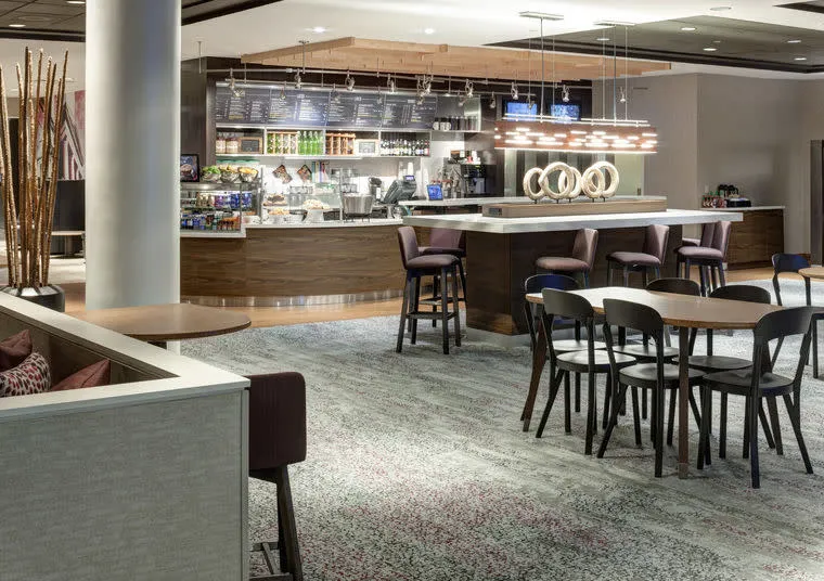A modern hotel café with a wood-accented counter, bar stools, and tables with chairs. The space features pendant lights, a display of snacks and drinks, and a carpeted floor with neutral tones.