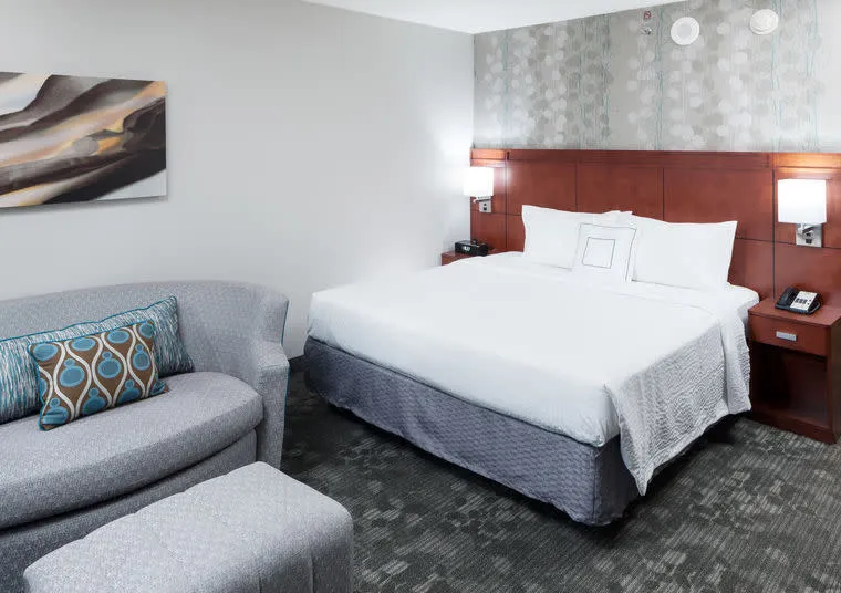 A modern hotel room with a large bed, white linens, wooden headboard, two bedside lamps, a grey patterned sofa with a cushion, an ottoman, carpeted floor, and abstract wall art.