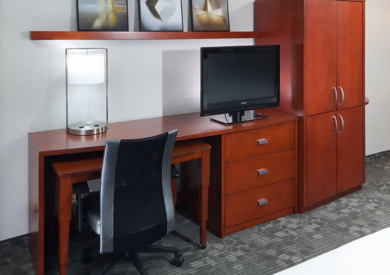A modern hotel room workspace with a wooden desk, black office chair, table lamp, flat-screen TV on a cabinet with drawers, and a tall matching wooden wardrobe. Artwork decorates a shelf above the desk.