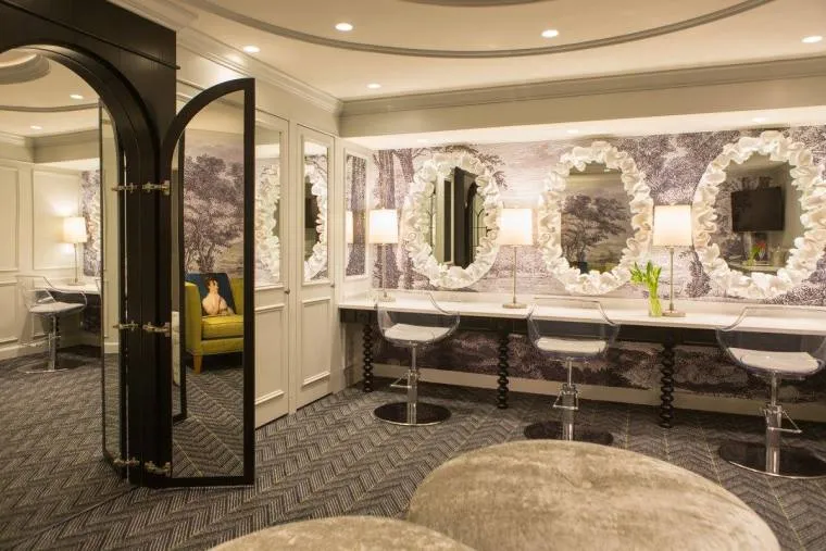Elegant dressing room with mirrored walls, three ornate oval mirrors above a long white counter with lamps, clear acrylic chairs, patterned carpet, and a seating area with plush stools. A yellow chair is visible in the background.