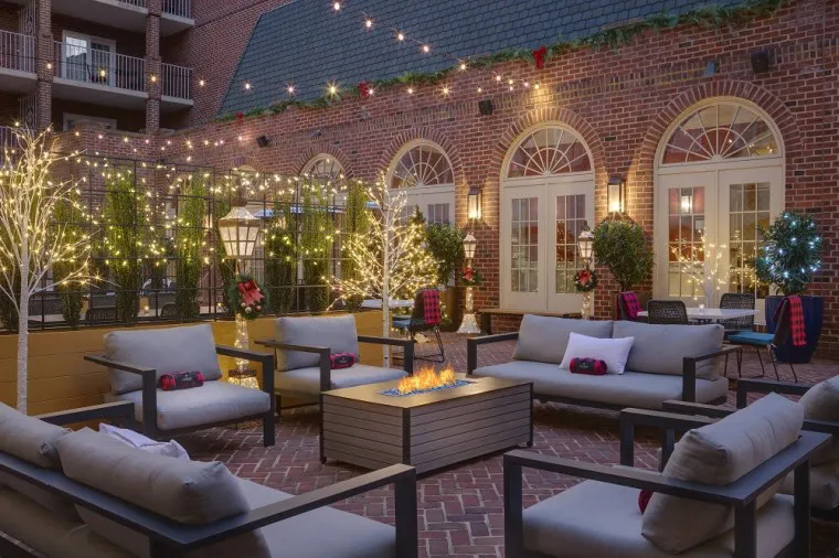 A cozy outdoor patio with cushioned seating around a fire pit, string lights and illuminated trees, set against a brick building with arched windows and greenery, creating a warm, inviting evening atmosphere.
